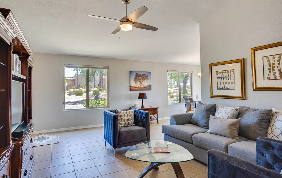 A comfortable living room featuring a sofa, accent chairs, a coffee table, artwork on the wall, and generous windows. The space is bright and filled with natural light, offering tranquil backyard views through the windows and a cozy setup for relaxing or socializing.