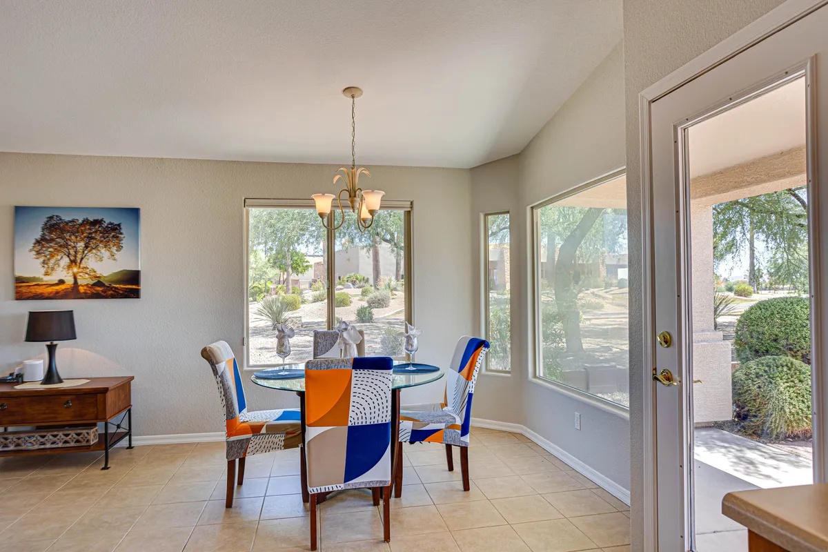 This image features a cheerful dining area with a large table, colorful chairs, expansive windows letting in abundant sunlight, and prominent artwork adorning the walls. The space exudes a social, lively vibe and suggests a place for community gatherings and shared activities.