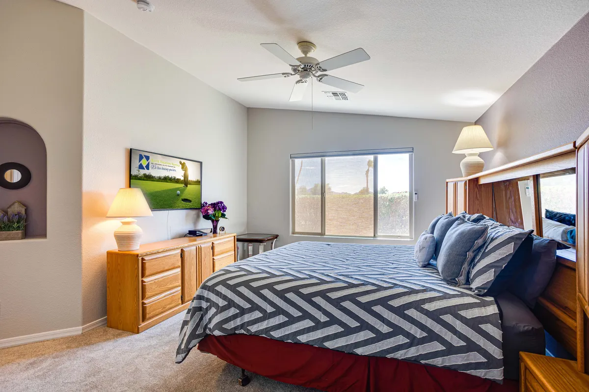 A serene bedroom featuring a plush bed with patterned bedding, a large window streaming natural light, a wooden dresser, and a bedside lamp create a restful, inviting atmosphere. The tidy layout and calm color scheme suggest this is the master suite.