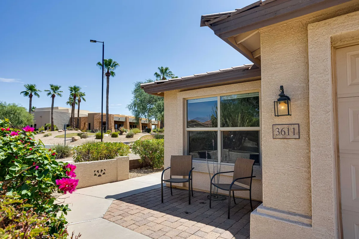 A wide view of a charming front porch framed by flowering plants and palm trees, with a visible house number and abundant greenery. The welcoming porch invites leisurely moments and social interaction, set in a vibrant neighborhood environment.